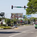 A wide palm-lined road with traffic lights, a green trolley bus and roadside billboards under a pale blue sky