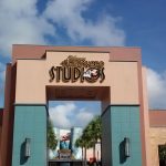A salmon pink archway with a teal base and a lit sign reading Hollywood Studios, with palm trees and a blue sketching-wizard hat beyond it