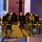 A brass and black multi-lens film camera rig hanging from a stand in a museum exhibit
