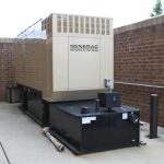 A tan standby generator enclosure sitting on a black fuel-tank base beside a brick wall