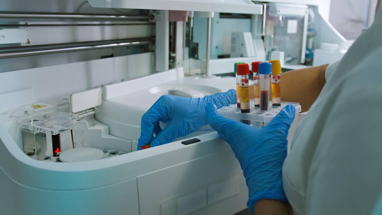 A technician in blue gloves loads capped blood tubes into a laboratory analyzer.