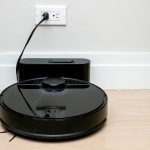 A black round robot vacuum resting against its charging dock beside a white wall with a power outlet