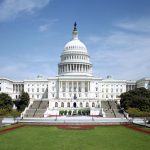 The west front of the United States Capitol under a blue sky, its white dome and steps seen straight on.