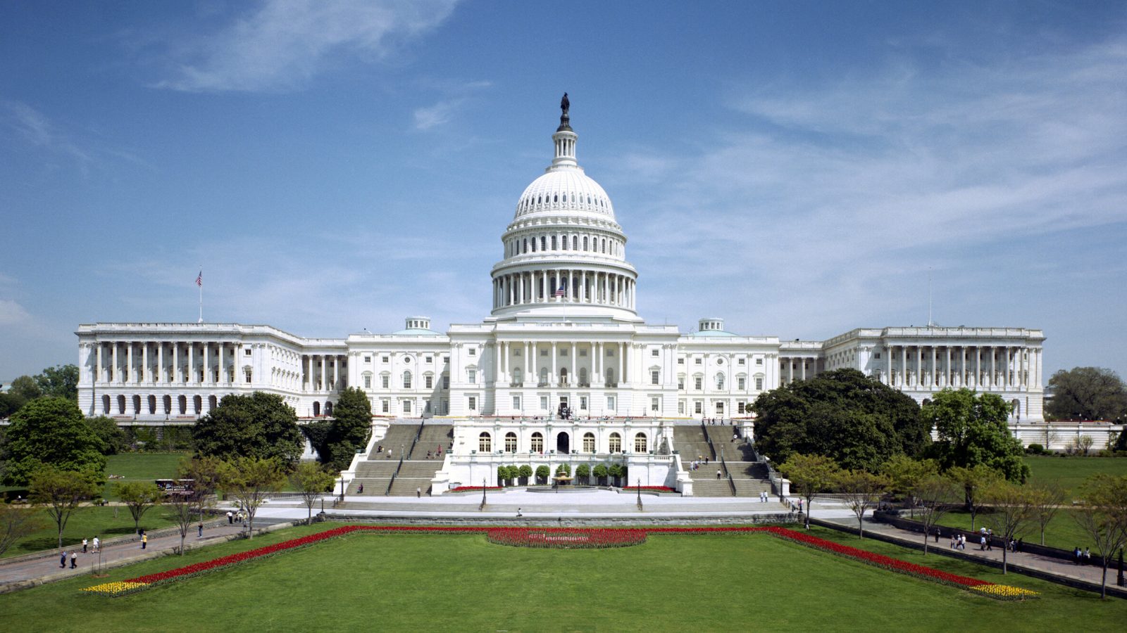 The west front of the United States Capitol under a blue sky, its white dome and steps seen straight on.