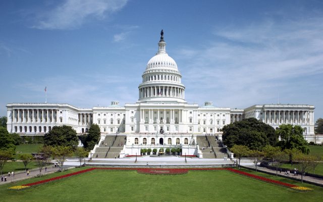 The west front of the United States Capitol under a blue sky, its white dome and steps seen straight on.
