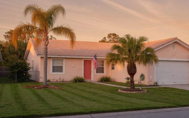 A modest single-story Central Florida home at golden hour with palm trees, a tidy lawn, and a small American flag by the front door.