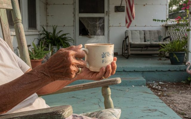The weathered hands of an older person holding a warm Florida coffee mug while seated on the porch of a modest home.