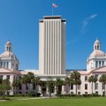 The Florida State Capitol in Tallahassee, its tall central tower flanked by the domed historic capitol building, under a clear blue sky.