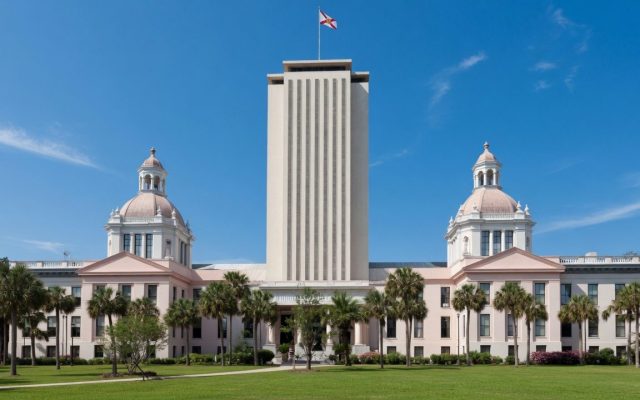 The Florida State Capitol in Tallahassee, its tall central tower flanked by the domed historic capitol building, under a clear blue sky.