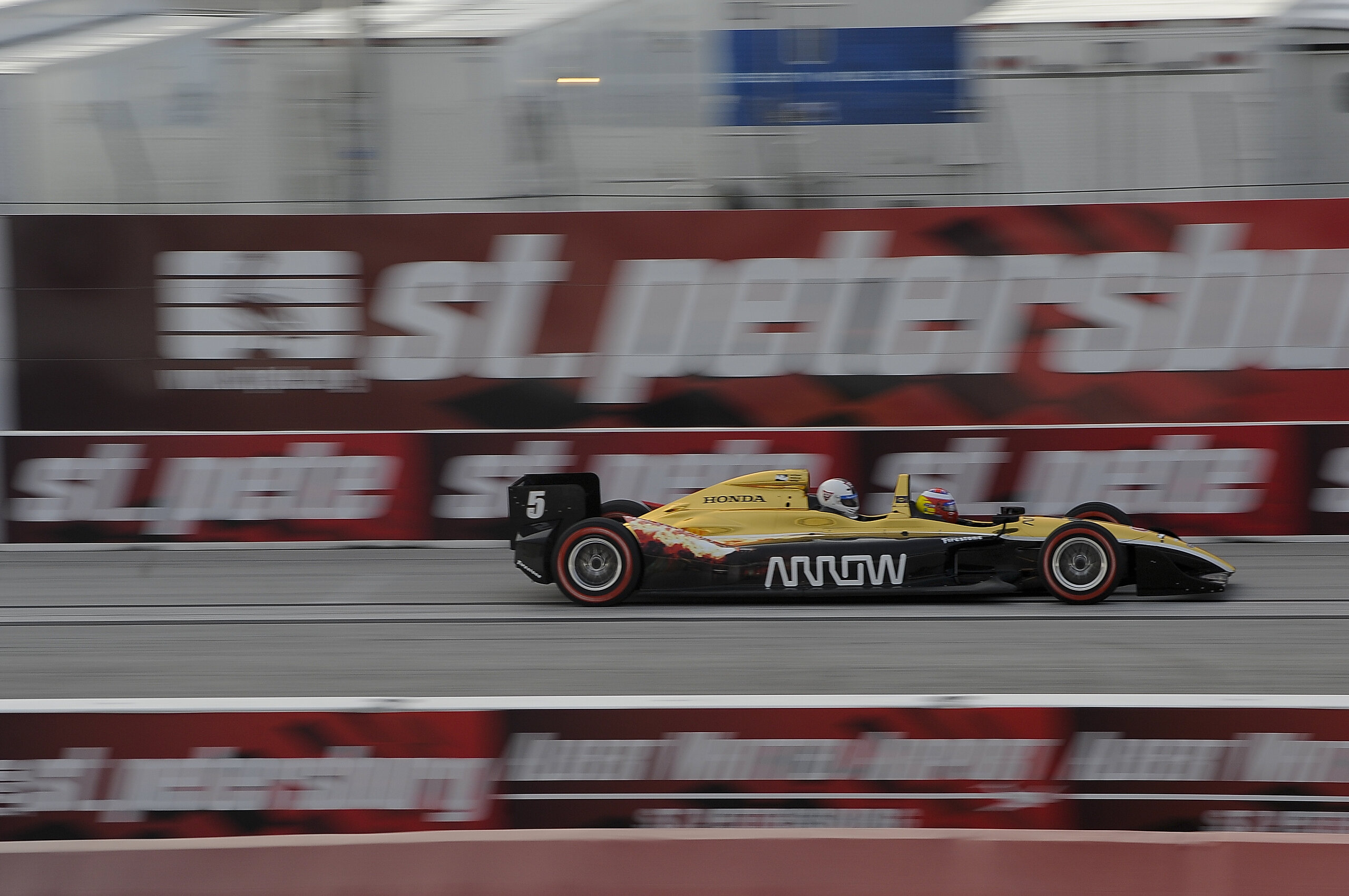 A number 5 open-wheel IndyCar at speed on a temporary street circuit, the barriers behind it blurred by motion.