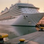 A large white cruise ship tied up alongside a quiet cruise terminal pier at sunrise, mooring lines and bollards in the foreground and calm water beyond