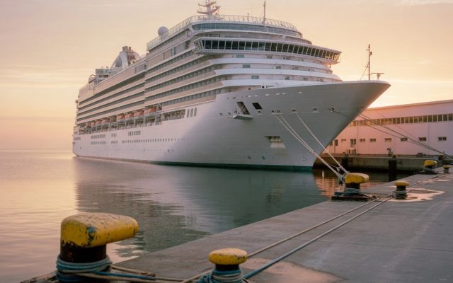 A large white cruise ship tied up alongside a quiet cruise terminal pier at sunrise, mooring lines and bollards in the foreground and calm water beyond