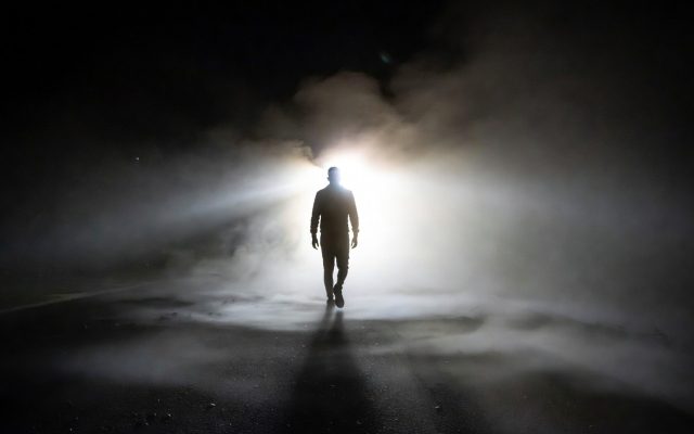 A lone figure walks through heavy fog toward a bright light on a dark road.