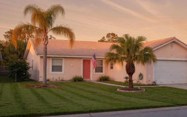 A modest single-story Central Florida home at golden hour with palm trees, a tidy lawn, and a small American flag by the front door.