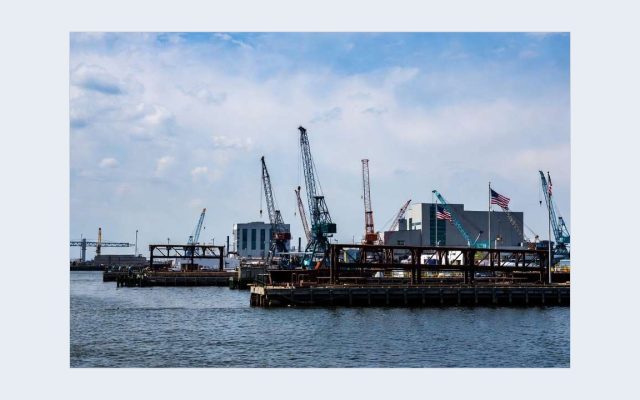 Waterfront view of the Philadelphia Navy Yard showing industrial cranes, piers, modern manufacturing buildings, and American flags flying under a blue sky