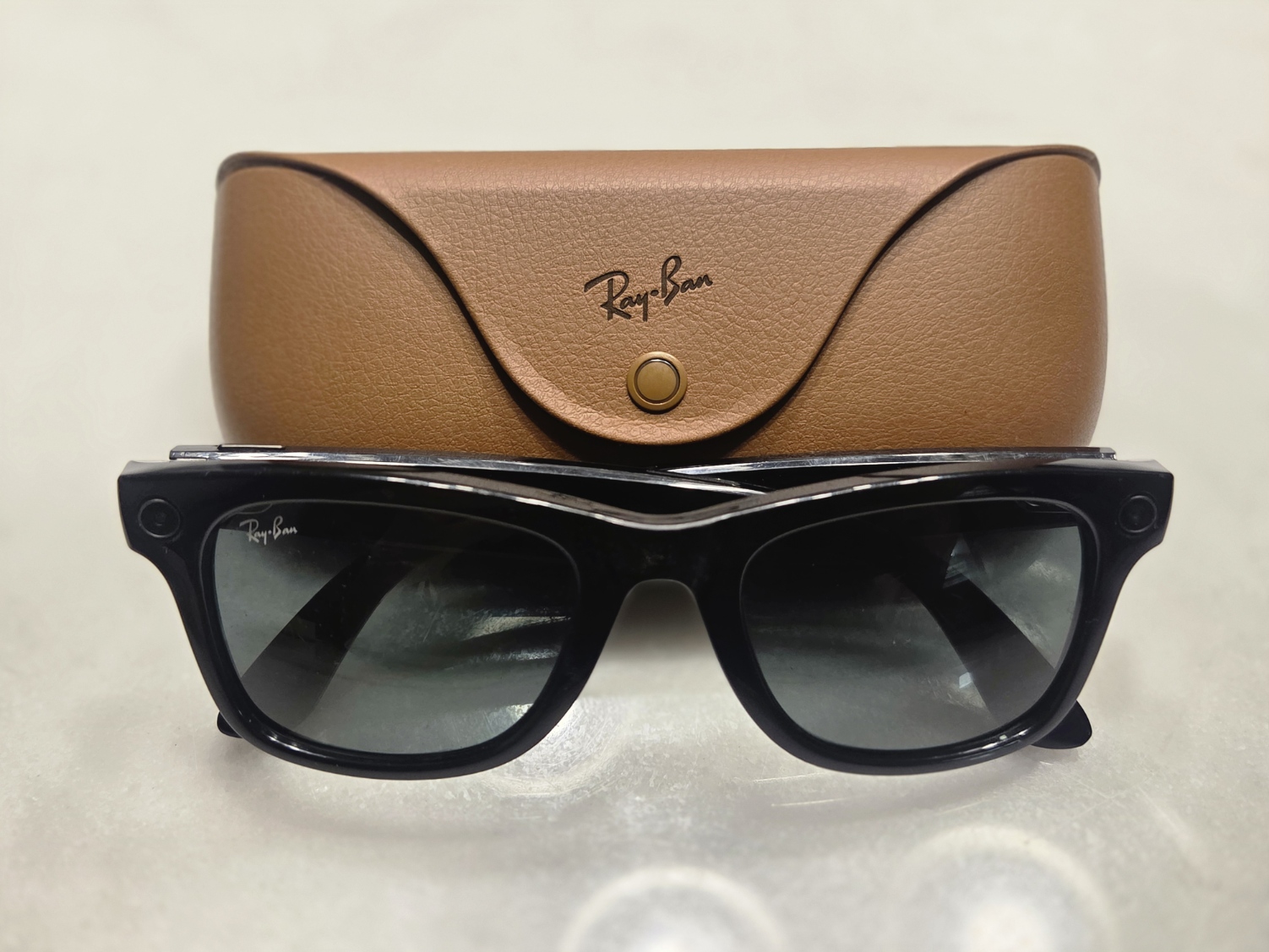 Black Ray-Ban Meta smart glasses with a small camera on the frame corner, resting in front of a brown leather case.