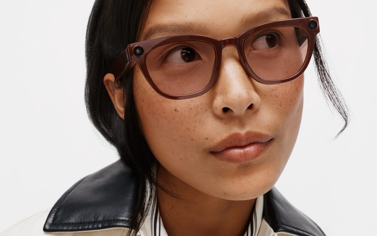 A person wears translucent brown smart glasses with a small camera lens set into the frame near the hinge.