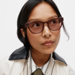 A person wears translucent brown smart glasses with a small camera lens set into the frame near the hinge.