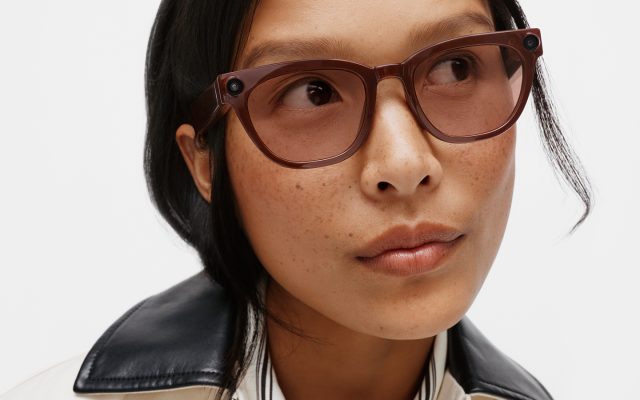 A person wears translucent brown smart glasses with a small camera lens set into the frame near the hinge.