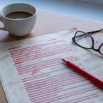 A printed page covered in red pen corrections on a desk beside reading glasses, a red pen and a cold cup of coffee