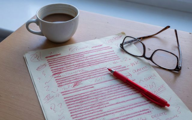 A printed page covered in red pen corrections on a desk beside reading glasses, a red pen and a cold cup of coffee