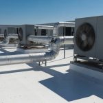 A flat commercial rooftop finished in bright white reflective coating under hard midday sun, with rooftop air-conditioning units and insulated ducting casting short shadows