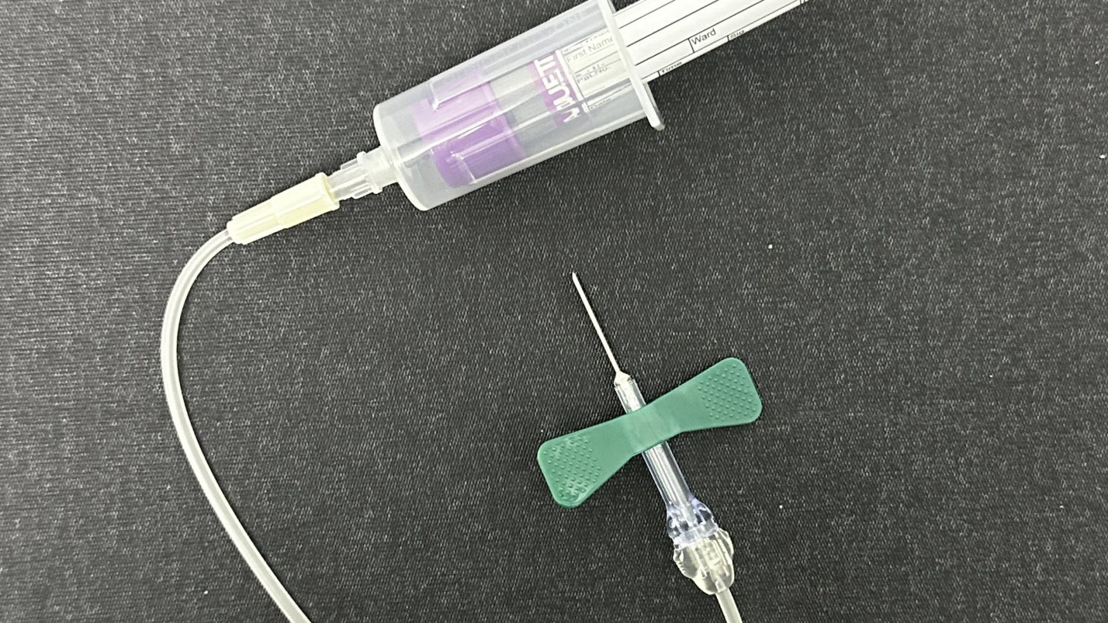A capped blood-collection tube beside a butterfly needle and tubing laid out on a dark cloth.