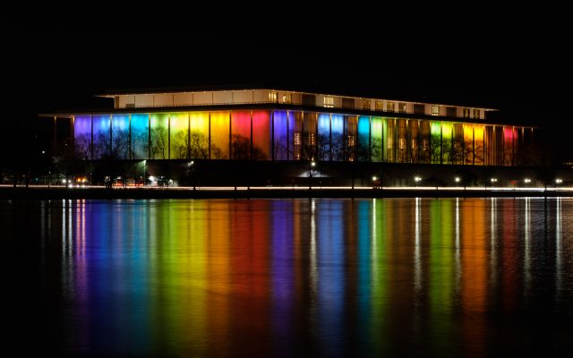 The John F. Kennedy Center for the Performing Arts at night, its colonnade lit in color and reflected in the Potomac