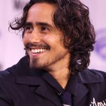 A smiling man with dark curly hair wearing a dark work shirt speaks on a stage