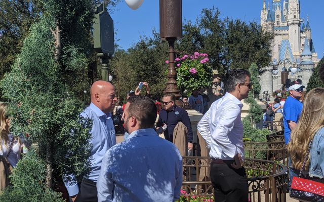 Bob Chapek, left, in a light blue shirt, talks with a man seen from behind while Josh D'Amaro, right, in a white shirt and sunglasses, stands nearby among guests, with Cinderella Castle in the background