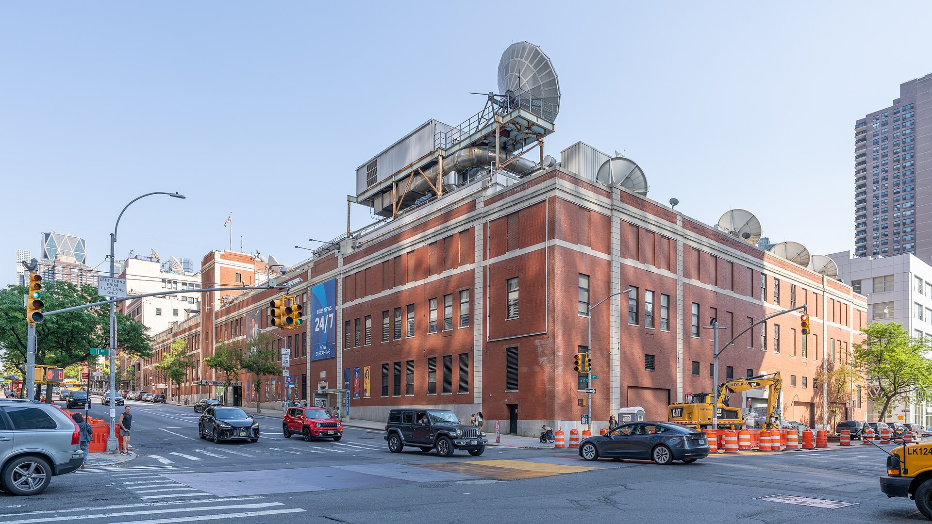 The red-brick CBS Broadcast Center on West 57th Street in Manhattan, with satellite dishes on the roof and a CBS News 24/7 banner, and traffic in the street