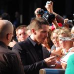 Christian Bale, in a dark suit, signs an autograph for fans holding cameras at the barrier on the red carpet in Leicester Square