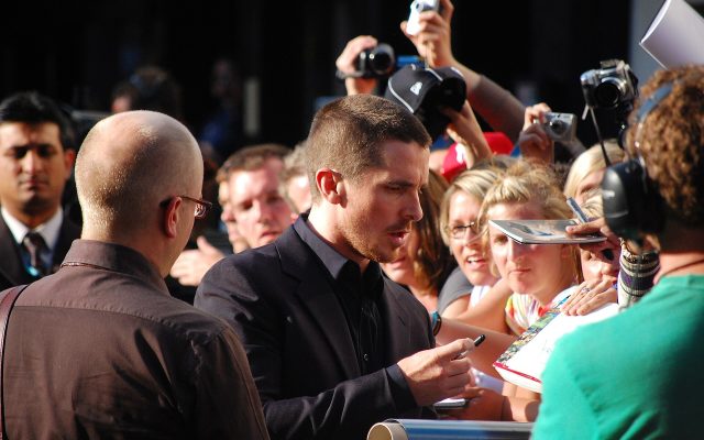 Christian Bale, in a dark suit, signs an autograph for fans holding cameras at the barrier on the red carpet in Leicester Square