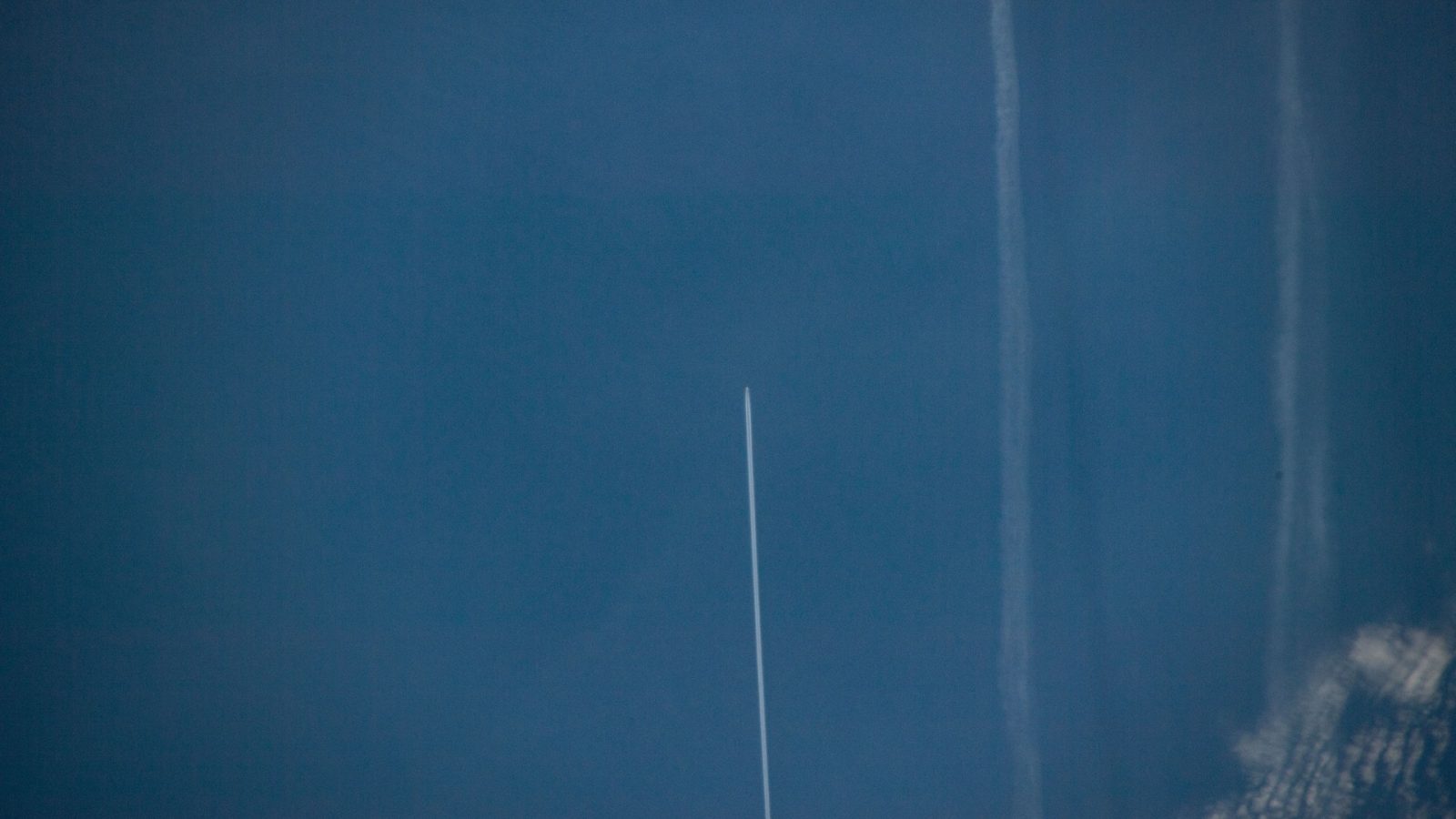 An aircraft and its fresh white contrail over the deep blue North Atlantic seen from the International Space Station, with older spread-out contrails nearby.