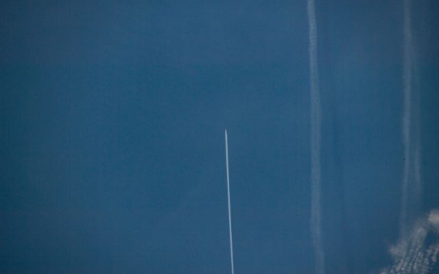 An aircraft and its fresh white contrail over the deep blue North Atlantic seen from the International Space Station, with older spread-out contrails nearby.