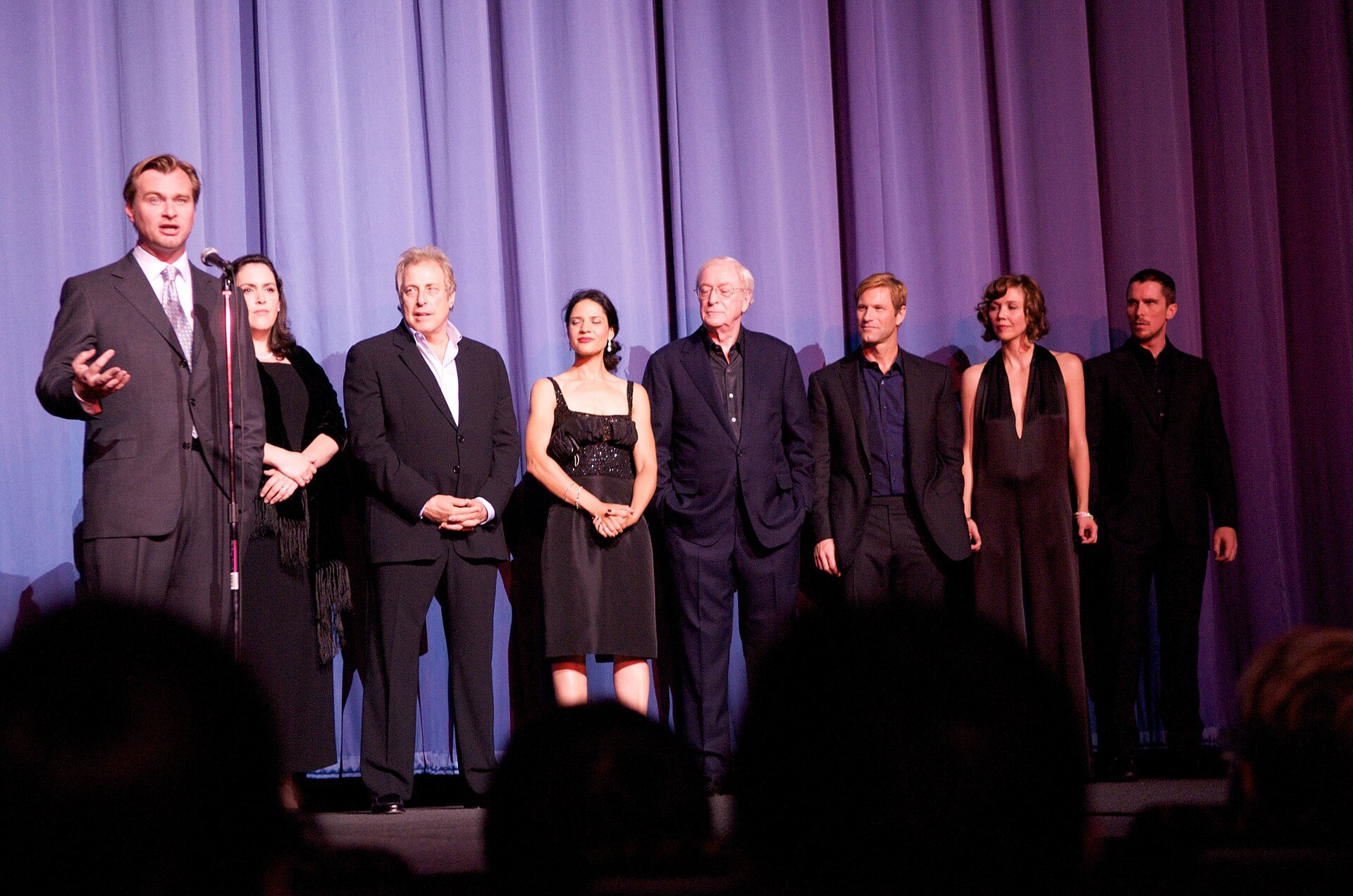 Christopher Nolan speaks at a microphone on a stage with a purple curtain, with seven members of the film's team standing in a line beside him and Christian Bale at the far right