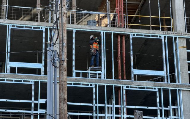 A hard-hatted worker balanced on the exposed steel framing of a large building under construction against an overcast sky.