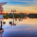 Colorful Disney Skyliner gondolas gliding over a still lake at dawn, mirrored in the water.