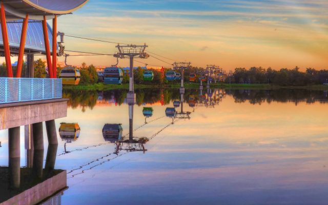 Colorful Disney Skyliner gondolas gliding over a still lake at dawn, mirrored in the water.