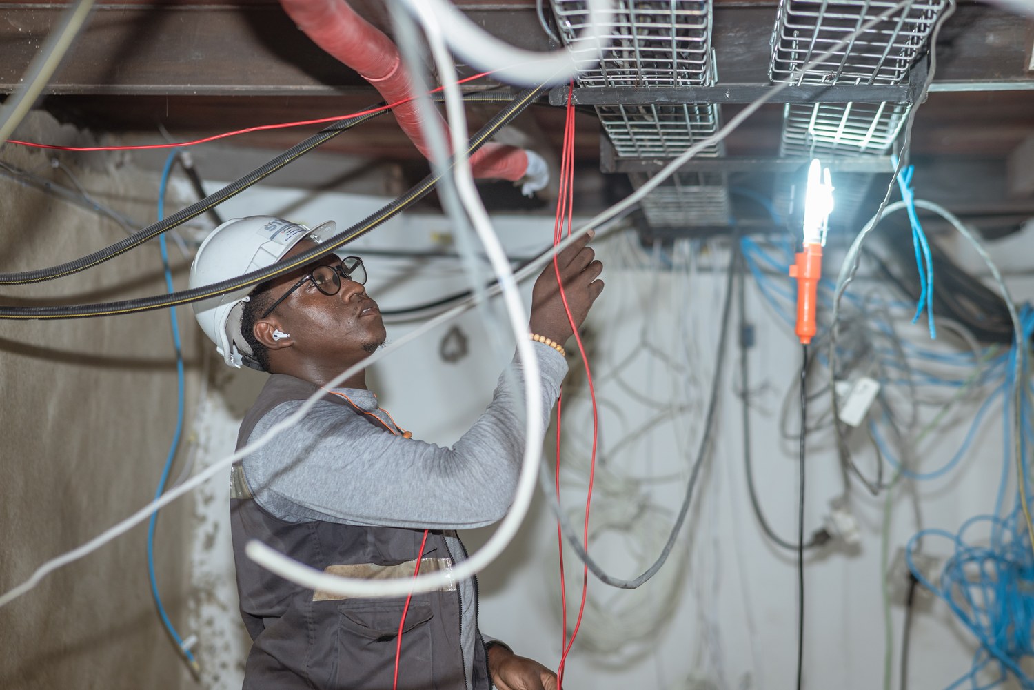An electrician in gloves works on a dense panel of colored wiring and breakers.