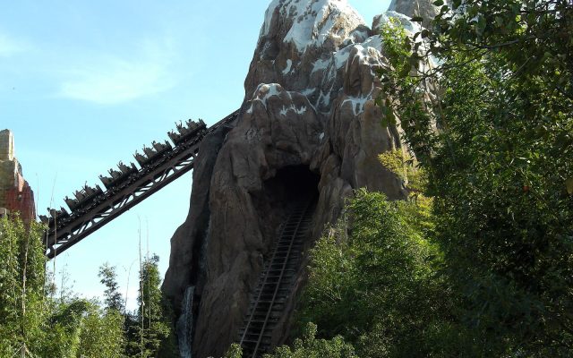 A roller coaster train full of riders seated two abreast climbs the lift hill beside the snow-capped artificial mountain at Expedition Everest, with a broken section of track running into a tunnel below.