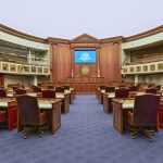 The empty Florida Senate chamber in Tallahassee, with rows of wooden desks and leather chairs facing the presiding officer's rostrum, where a screen reads Welcome to the Florida Senate.