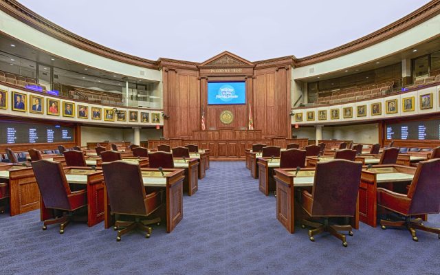 The empty Florida Senate chamber in Tallahassee, with rows of wooden desks and leather chairs facing the presiding officer's rostrum, where a screen reads Welcome to the Florida Senate.