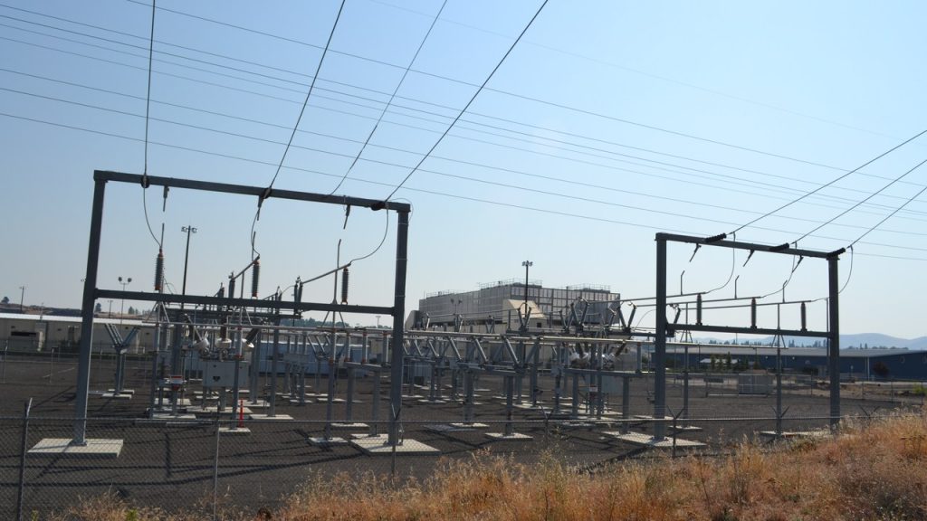 An electrical substation with steel towers and power lines in front of Google's data center buildings at The Dalles, Oregon, under a clear sky