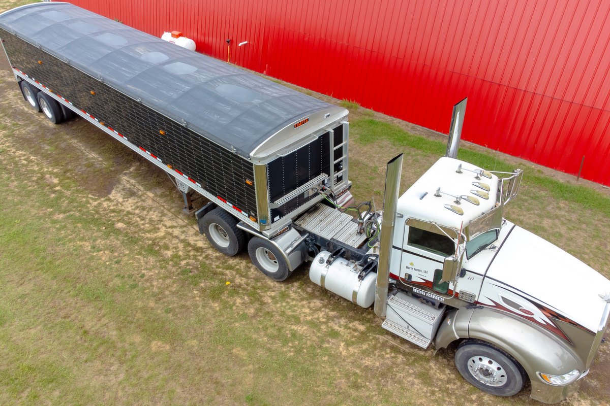 A semi truck with a grain hopper trailer parked beside a barn.