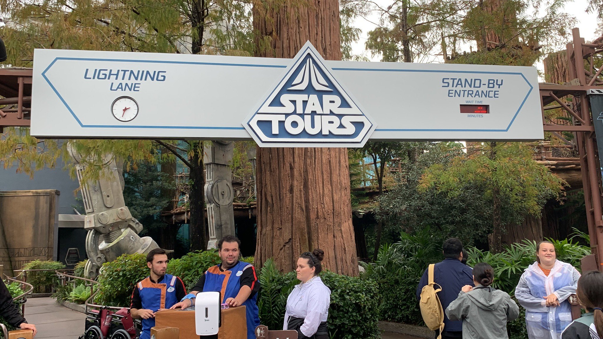 An overhead Star Tours sign at Disney's Hollywood Studios marks the Lightning Lane on the left, with a clock face, and the Stand-By Entrance on the right, with a wait-time display, above two cast members at a podium and waiting guests