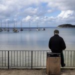 An older man in a dark jacket sits alone on a post by a railing, looking out over a calm bay dotted with sailboats.