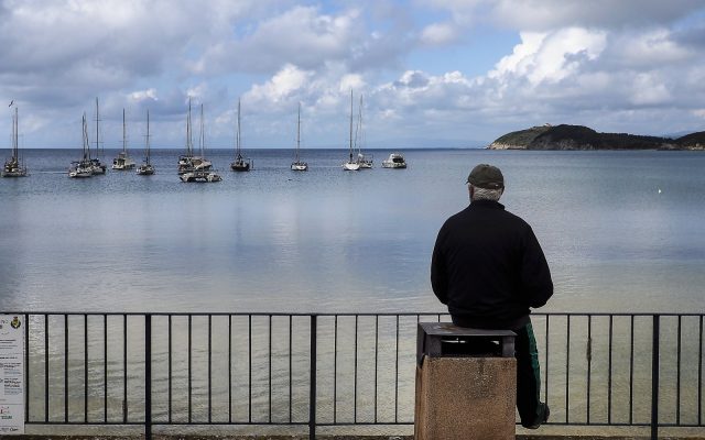An older man in a dark jacket sits alone on a post by a railing, looking out over a calm bay dotted with sailboats.