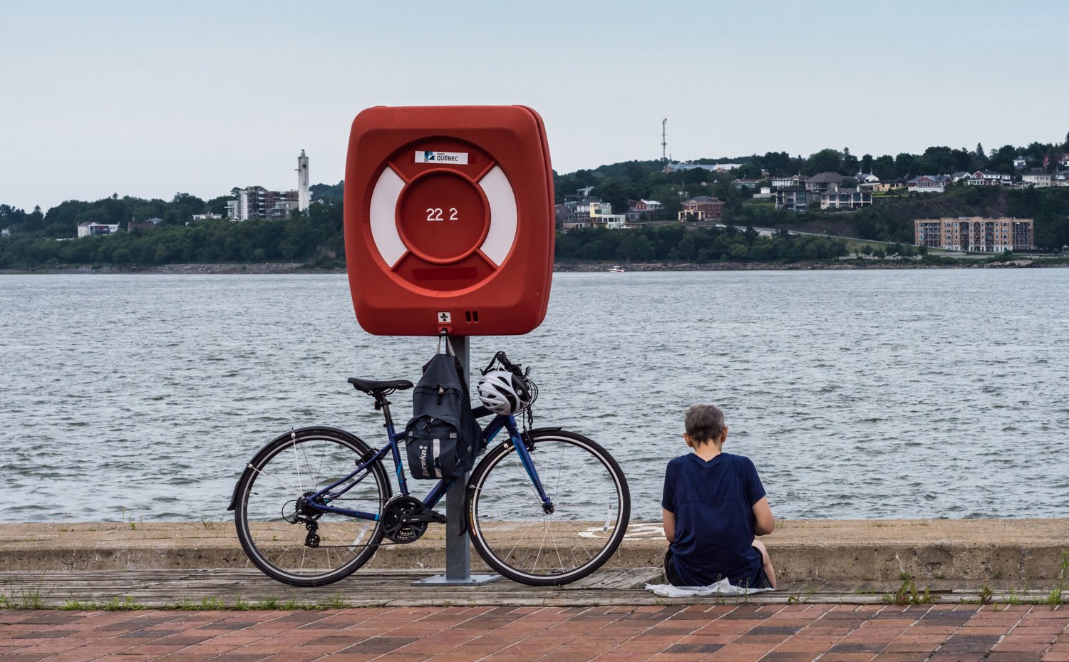 A small solitary figure sits at the edge of a stone quay beside a bicycle and a red lifebuoy, a town visible across the water.