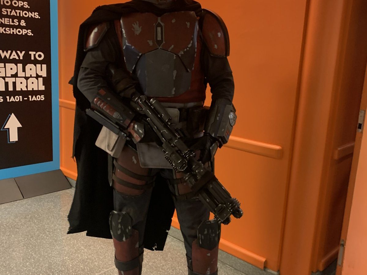 A full-armor Mandalorian cosplayer in beskar helmet and cape holding a blaster rifle at a convention.